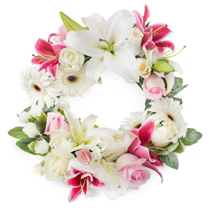 This image shows an elegant, fully open circular funeral wreath, carefully arranged against a crisp white background, like it has just been completed on a florist's workbench in Queensbury. The design is rich with white, blush pink, and deep fuchsia tones, all balanced to create a feeling of composure and reverence. Dominating the wreath are large white lilies, their broad petals sweeping outward in graceful arcs, each bloom revealing vivid green pistils and golden-tipped stamens that catch the light. Interspersed among them are bold fuchsia and deep magenta lilies, easily recognized by their star-shaped forms and delicate speckling, which bring a sophisticated splash of colour without overwhelming the serenity of the piece. Between these feature blooms sit clusters of soft pink roses and creamy white roses, some tightly closed and others half open, the differing stages of bloom symbolizing the span of a life and the memories left behind. White gerbera daisies punctuate the circle, their layered petals and dark central disks providing both texture and gentle contrast, almost like small, attentive eyes. Smaller white filler flowers, probably miniature carnations or spray chrysanthemums, nestle into every gap, giving the arrangement fullness and a soft, cloud-like density. Around the entire ring, glossy green foliage frames the flowers, overlapping leaves creating depth and reinforcing the perfect circular shape that suggests eternity and unending love. This thoughtfully designed wreath feels ideal for a funeral or memorial service, the kind a Queensbury family might send to a service near Queensbury Park or along Kingsbury Road, conveying honour, remembrance, and quiet support through its peaceful colour palette and meticulous craftsmanship.