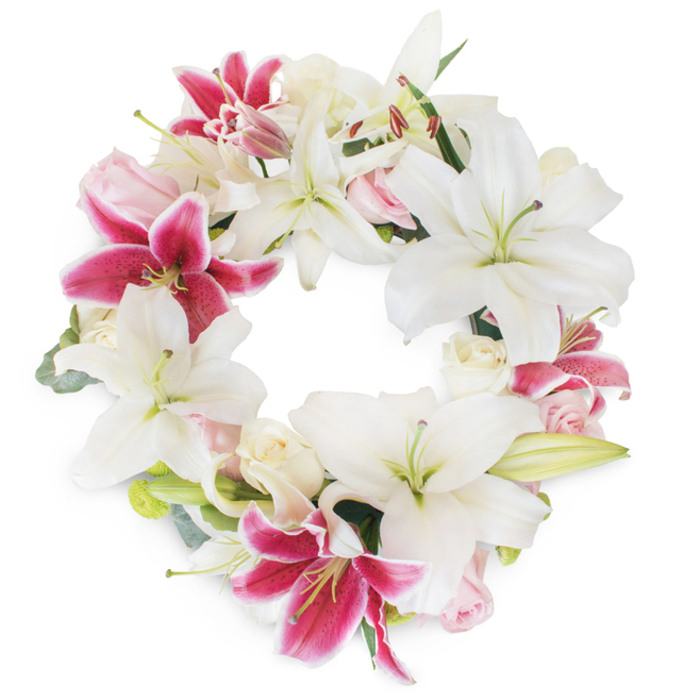 Against a crisp white backdrop, a lavishly designed floral wreath forms a radiant circle-its centre left open, as if inviting thoughts of peace and celebration. This composition showcases the elegant interplay between large, pristine white Oriental lilies and dazzling fuchsia-pink Stargazer lilies, their petals star-shaped and luminous, some gently curling outward to reveal pale green stamens and the bright, sunlit heart of each bloom. The deep magenta hues and delicate speckling of the fuchsia lilies provide vivid focal points that catch the eye and enliven the overall design. Between these show-stopping blossoms, soft blush-pink and ivory white roses nestle, their forms varying from tightly furled buds to fully unfurled petals, layering the arrangement with romantic softness and refined texture. Accents of silvery-green foliage-perhaps sprigs of eucalyptus or slender lily leaves-emerge from the lush bed of flowers, supplying structure and a subtle woodland fragrance that would be familiar to those strolling through Queensbury's parks. The arrangement is perfectly symmetrical, each bloom carefully placed to symbolise unity, eternal life, and reverent remembrance. This wreath, created with exceptional care and understanding of local tradition, is ideal for funeral services or commemorative occasions throughout Queensbury, capturing both the dignified sorrow and the enduring affection that shape each tribute.