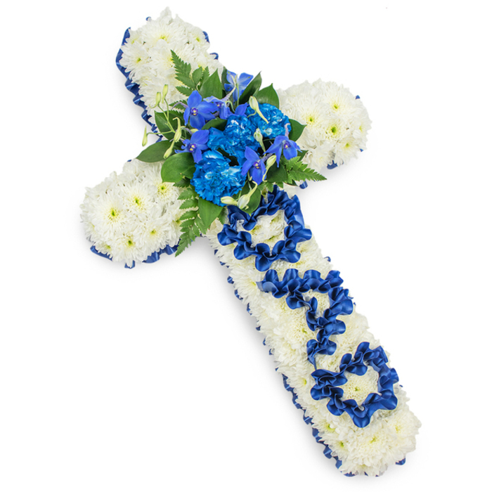 This image shows a large, cross-shaped funeral tribute set against a crisp white background, allowing every floral detail to stand out clearly. The entire body of the cross is densely covered with pristine white spray chrysanthemums, their small, rounded blooms tightly massed together to form a smooth, cushioned surface that looks both soft and substantial. Gently curved petals overlap to create a sense of fullness, suggesting comfort and purity, like a quiet blanket of snow. Around the outer edge of the cross, a wide band of royal blue satin ribbon has been meticulously pinned and folded, outlining the form with a rich, jewel-toned frame. The ribbon's subtle shine catches the light, adding a sense of formality and reverence to the tribute. At the central junction of the cross, where horizontal and vertical arms meet, a bold cluster of royal blue flowers draws the eye: generous, ruffled heads of blue carnations are nestled together with more slender, upright blue blooms that resemble delphiniums or orchids, adding delicate spikes of colour and structure. Dark green fern fronds and layered foliage weave between the blue flowers, offering contrast and evoking the calm of a shaded garden or a mature tree line you might see walking through the quieter corners of Queensbury. Running down the vertical arm, the word DAD is clearly visible, crafted from loops and neatly folded lengths of the same blue satin ribbon, standing out against the white chrysanthemum base. This carefully made detail makes the piece unmistakably personal, a sincere memorial for a father at a funeral, wake, or graveside ceremony in the local community. The overall impression is solemn yet tender, a crafted symbol of faith, remembrance, and enduring love.