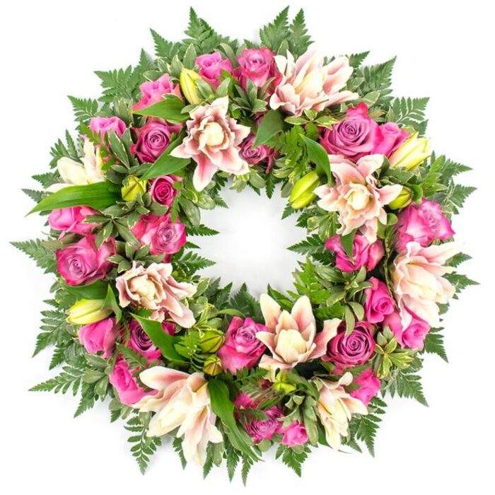 A meticulously crafted circular funeral wreath dominates the frame, set against a seamless white background that throws every flower, leaf, and shadow into gentle relief. The ring is fully covered, with no gaps in the greenery, giving a sense of completeness and continuity. At its heart are sumptuous roses in vivid fuchsia, rich magenta, and softer blush pink, distributed evenly around the circle so that the eye moves smoothly from one bloom to the next. Some roses are tightly furled, with pointed centres and smooth outer petals, while others are open and layered, revealing a complex swirl of velvety texture. Interlaced among the roses are generous double lilies, reminiscent of garden hybrids like soft-pink stargazers, but rendered here in pastel tones of cream, blush, and pale peach. Their multiple petal layers create a frilled, almost cloud-like effect, and in a few blooms the deeper pink veining towards the tips is just visible. Upright lily buds in bright, citrus green emerge at intervals, suggesting ongoing life and the promise of further flowering. The entire composition rests on a luxurious base of foliage: dark green fern fronds trace the curves of the wreath, their fine leaflets adding delicacy, while shiny, broader leaves and occasional variegated accents build depth and volume. The interplay between the cool greens and warm pinks feels balanced and serene. This floral ring would be equally at home on an easel stand during a crematorium service near Queensbury or laid flat during a quieter family farewell. Crafted with the care you would expect from a dedicated local team at Flower Delivery Queensbury, it offers a refined, traditional expression of sympathy, remembrance, and respect.