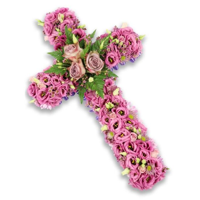 A beautifully composed floral cross is displayed against a stark white backdrop, the crisp background highlighting every curve and contour of the flower-covered shape. The cross itself is fully enrobed in layers of lisianthus flowers, their petals softly fluted and slightly curled, in a palette of delicate pinks, mauves, and light purples that blend into one another like a gentle evening sky over Queensbury. The densely packed blossoms make the cross appear plush and three-dimensional, giving the sense that you could reach out and feel the cool, velvety texture of each bloom. At the central junction, three prominent roses in dusty rose and muted mauve tones have been carefully positioned to form a calm, focal cluster, their open faces turned slightly outward as if in quiet blessing. Framing these roses are light, airy fern fronds and compact green leaves, providing a fresh, woodland contrast that lifts the pastel colours and adds depth to the design. Dotted throughout the cross are delicate filler flowers in shades of pink and lilac, along with a few pale cream or white blossoms with tiny centers, and occasional soft green buds yet to fully open, suggesting ongoing life and renewal. Along the edges of the vertical and horizontal arms, slender bands or hints of deep purple ribbon reveal themselves between blossoms, outlining the cross without disturbing its softness. Created with the care of a seasoned florist at Flower Delivery Queensbury, this arrangement feels deeply suitable for a funeral or memorial in the local churches and crematoria, offering a dignified symbol of faith, remembrance, and gentle, enduring love.