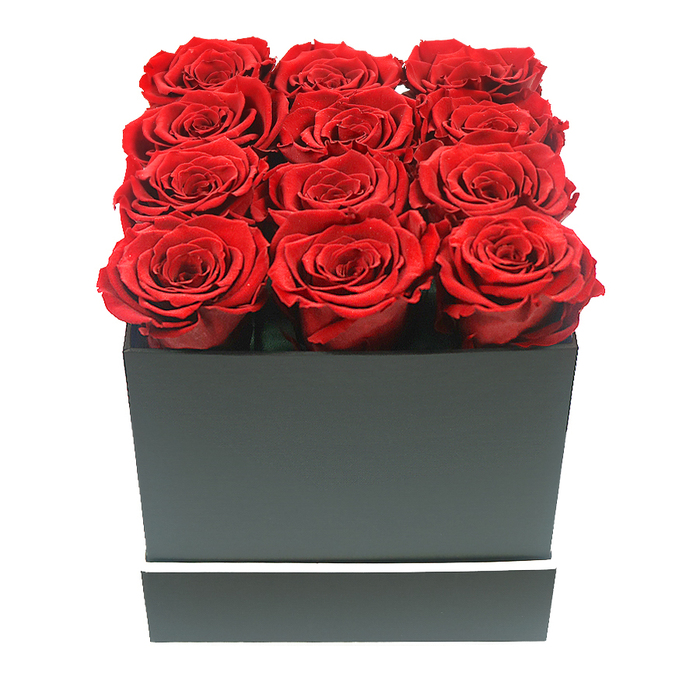This image shows a stylish, tightly framed composition of a red rose hatbox arrangement that could easily be imagined arriving from Flower Delivery Queensbury to a chic home just off Queensbury Circle. At the centre are twelve sumptuous roses in a rich, ruby red shade, each bloom fully open and perfectly rounded, so their velvety petals overlap like layers of silk. The flowers are arranged with mathematical precision in a 3x4 grid, filling every corner of the square opening and extending slightly above the rim so that the tops of the roses form a plush, level cushion of colour. Each rose head appears evenly sized and at the same stage of bloom, evoking the look of preserved or 'eternal' roses chosen for maximum uniformity and longevity. The container is a matte black square box with crisp, neat edges and a calm, modern feel. Its surface is smooth and untextured, absorbing light and allowing the crimson blooms to stand out. Around the lower part of the box runs a fine, bright white band that wraps around horizontally, a subtle design feature that lends a contemporary, almost designer-gift quality. The background is a clean, seamless white, with no extra décor or props, focusing attention solely on the contrast between the dark box and the glowing red flowers. Soft, even lighting casts delicate shadows along the petals and edges of the box, giving a gentle sense of depth without overpowering the arrangement. This luxurious hatbox of roses would be perfect for a Valentine's Day delivery to a restaurant near Queensbury underground station, a significant anniversary in a flat on Honeypot Lane, or any occasion where a client in Queensbury wants to send a romantic, long-lasting floral keepsake with a distinctly modern look.