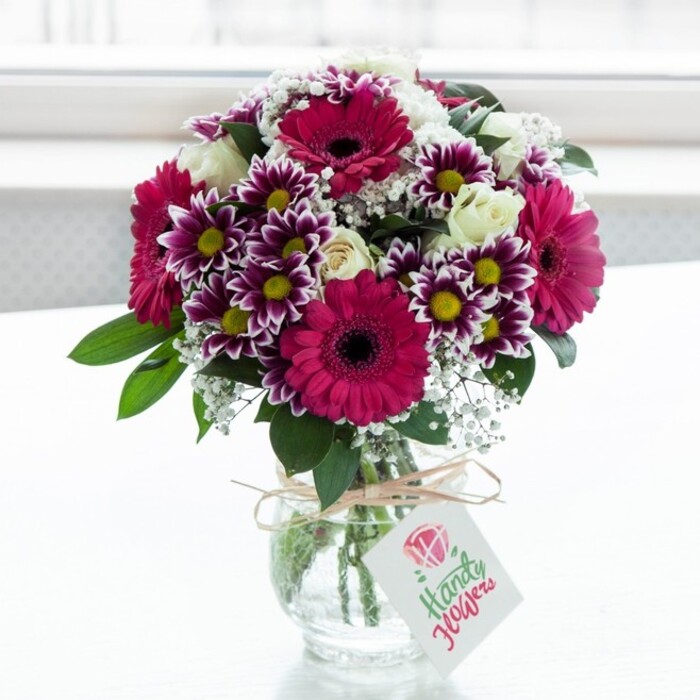 A beautifully composed mixed flower bouquet sits at the centre of the frame in a clear, slightly rounded glass vase, set against a luminous white backdrop that suggests a sunlit windowsill in a tidy Queensbury home. The arrangement is a celebration of pink and purple tones, anchored by several striking gerbera daisies in vivid fuchsia and magenta, their broad, symmetrical petals radiating around deep, dark central discs that create a bold, graphic contrast. Nestled between these statement blooms are generous clusters of two-tone chrysanthemums, each petal edged in rich purple that gradually softens to white near the bright yellow centres, giving the bouquet a lively, almost painterly texture. Creamy white spray roses are scattered thoughtfully throughout, some fully open with layered, cupped petals, others still in bud form, adding both elegance and a sense of unfolding time. Fine sprays of white gypsophila, with their myriad tiny blossoms, are threaded delicately between the larger flowers, creating a soft halo effect and giving the arrangement an airy, romantic quality reminiscent of blossom trees along local streets near Queensbury Circle. Lush dark green foliage frames and supports the blooms, their slightly glossy surfaces catching the light and enhancing the sense of freshness. The clear glass vase reveals neatly cut stems splayed in the water, and around the neck a length of natural twine is tied into a simple, rustic bow, conveying a crafted, hand-finished touch. A small white gift tag dangles from the twine, adorned with a stylised pink rose graphic and understated script, hinting at a heartfelt message rather than any bold branding. The soft, even lighting picks out the nuances in each petal and leaf, making the arrangement feel freshly prepared and ideal for same-day gifting within Queensbury, whether for birthdays, get-well wishes, or to bring a warm burst of colour to a quiet living room.
