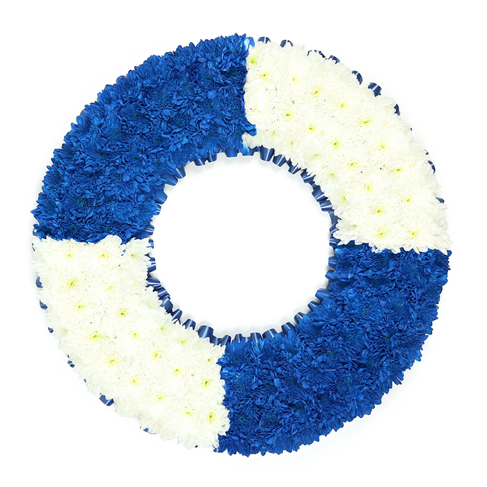 A beautifully composed maritime funeral wreath takes centre stage against a spotless white background, capturing the eye with its strong circular form and carefully balanced colour. Crafted to mirror a traditional lifebuoy, the ring is built entirely from tightly clustered, small-flowered blooms, most likely chrysanthemums chosen for their longevity and steady shape. The circle is crisply divided into four quarters: starting from the top left and moving clockwise, the sections alternate between a saturated royal blue and a pure, bright white. The blue areas are so rich and even that they almost appear like soft velvet from a distance, while the white segments glow with a quiet, gentle clarity, suggesting innocence and peace. Each flower head is pressed firmly into place, the petals overlapping and interlocking so there is no visible foliage, stem, or gap-just an unbroken surface of colour that follows the curve of the ring. Along the inner edge, just inside the flowers, a slim band of dark blue ribbon is delicately ruffled, forming a neat, continuous trim that deepens the nautical character and emphasises the hollow centre of the wreath. The background is completely uncluttered, ensuring that every detail of the craftsmanship stands out: the precision of the quadrants, the smoothness of the circular outline, and the harmony between the colours. This type of tribute would feel especially meaningful at a service for someone from Queensbury with links to maritime work, naval service, or perhaps a cherished family member known for their protective, dependable nature. It speaks softly of safe passage, of being held and watched over, and of a final journey taken with respect and love.