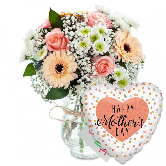 This image presents a lovingly composed Mother's Day gift set, photographed against a crisp white background that keeps every detail clear and uncluttered, much like a bright morning in Queensbury. On the left stands a rounded bouquet of mixed spring flowers, arranged in a transparent glass vase with smooth sides and a casual loop of light brown twine tied neatly around its neck. The bouquet itself is lush and full, dominated by several soft peach and blush-pink roses, their petals gently unfurling to show velvety spirals and warm, romantic tones. Nestled among them are striking peach gerbera daisies with dark brown centres, their flat, sunburst petals bringing a cheerful, modern accent that draws the eye. Between the larger blooms, clusters of white baby's breath weave through like light, airy clouds, while small white chrysanthemums with subtle pale green centres add texture and volume, giving the bouquet a rich, layered look. Shades of fresh green foliage frame and support the flowers, hinting at hedgerows and gardens around Queensbury Park, and making the pastel colours glow more intensely. In front of the vase, slightly to the right and partly overlapping the glass, a heart-shaped foil balloon leans forward, as if nudging into the scene. The balloon is pure white and generously dotted with sparkling gold and silver circles that mimic confetti, creating a party-ready feel. At its centre, a bold peach heart encloses the message HAPPY Mother's DAY in contrasting black lettering, combining a strong sans-serif style for HAPPY with an elegant, handwritten script for Mother's Day. A small white greeting card peeks discreetly between the vase and balloon, reinforcing the idea of a complete gift. The overall mood suggests a fresh, ready-to-deliver surprise for a special mum in Queensbury.