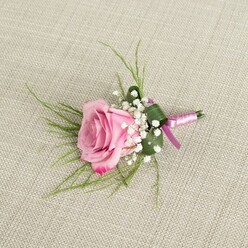 An elegant pink rose boutonniere rests on a natural, light beige linen surface, its romantic simplicity evoking a Queensbury ceremony just off Victoria Road or a prom evening beginning with nervous smiles and careful dressing. The single rose is captured from above, half-open and perfectly poised between bud and full bloom, with petals that transition from a soft, powdery blush at the centre to a richer, almost raspberry pink at the outer edges. Each petal appears velvety and fresh, gently curved and layered to create a sense of depth and softness. Surrounding the rose, airy sprays of white baby's breath add a delicate, cloud-like frame, their tiny star-shaped blossoms clustered closely to highlight the larger central flower. Wisps of slim, fresh green foliage extend lightly from the base, giving the arrangement a slightly playful lift, while a broader, darker green leaf is folded snugly around the stems to provide structure and a sense of polished design. The stems are carefully wrapped in a satin ribbon that shimmers with tones of lavender-pink and deeper magenta, its smooth texture contrasting with the more organic weave of the linen beneath. Natural, diffused light glides across the scene, emphasising the rose's gentle colour gradients and the intricate textures of petal, leaf, and fabric, while subtle shadows define the boutonniere's compact, handcrafted form. This floral detail, reminiscent of the work you might collect from a trusted Queensbury florist before walking to a nearby venue, feels both timeless and contemporary, perfectly suited to a groom, groomsman, or prom date wanting a refined touch of romance on their lapel.