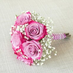 Bathed in gentle daylight across a light beige linen reminiscent of homes and venues near Queensbury's Kingsbury Road, this radiant bridal bouquet celebrates both floral artistry and local connection. The bouquet's rounded composition brings together five remarkable roses, each one displaying a mesmerising dual-tone effect: their innermost petals reveal soft washes of lavender and pink, gracefully intensifying to vibrant magenta and fuchsia along the outer ruffles. Tucked between these statement blooms, clouds of pure white gypsophila-baby's breath-drift delicately, introducing a caress of softness and textural contrast that echoes the blossoms of local gardens in early summer. The tactile pleasure is heightened by the meticulously wrapped, deep purple satin ribbon handle, adorned in a harmonious spiral of iridescent pearl strands. This refined accent evokes a sense of occasion, reminiscent of formal celebrations at Queensbury's cherished community halls. Beneath, the natural green-brown stems remain on show, connecting this elegant display to the earth and authenticity that defines local craftsmanship. The bouquet radiates harmony and quiet joy, ideal for a wedding aisle or special vow exchange, carrying an air of both innocence and sophistication. Its thoughtful composition and luxurious accent details reflect a deep understanding of local style, making it not just a bouquet, but a treasured keepsake of a memorable Queensbury celebration.