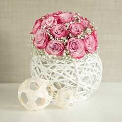 A delicately styled floral still life features a dome of pale pink roses and airy white baby's breath rising from a sculptural white basket-style vase, as if prepared by a careful florist just off Queensbury Station. The roses form a near-perfect sphere, their petals unfolding from soft, shell-pink hearts into deeper, more saturated pink edges, with a few blooms leaning slightly outward to create a natural, garden-picked feel. Between these roses, countless tiny blossoms of Gypsophila weave through the gaps, their crisp white colour and fine stems adding a misty, almost bridal veil effect around the pink heads. The bouquet is cradled in a rounded container made from narrow white strands, woven into an open lattice that brings to mind modern wicker; the spaces between the strands reveal hints of shadow, giving the vase depth and a crafted, artisan look. Resting on the clean white tabletop in front and to the side are two decorative spheres of different sizes, made from pale, interwoven wooden strips. Their loose, geometric patterns quietly echo the structure of the vase, bringing textural harmony to the composition. Behind, a softly textured wall in gentle beige or cream provides an unobtrusive backdrop, similar to the calm interiors of Queensbury family lounges and dining rooms. Light falls evenly across the arrangement, emphasising the roses' velvety petals and the airy construction of both the vase and the wooden orbs. The overall impression is of calm celebration and understated romance, perfect for a birthday breakfast table, a low-key engagement toast, or a thoughtful floral delivery to a loved one living along Honeypot Lane or nearby streets.