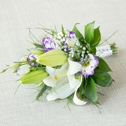 An elongated hand-tied bouquet of white lilies and purple lisianthus lies on a pale beige, subtly woven fabric that looks like softly lit linen, angled diagonally as though it has been carefully placed on a dressing table before a ceremony. At the heart of the arrangement, generous white Oriental lilies unfold their velvety petals, each bloom perfectly open to reveal tall yellow-orange stamens and fine speckles of rust along the inner curves, giving the flowers depth and character. Alongside these stars of the bouquet, several light green lily buds stand upright and slightly tapered, still tightly closed but clearly on the verge of opening, adding a sense of anticipation. Between the lilies, lisianthus flowers weave through in shades of white and purple: some petals are creamy with a vivid violet edge, others drift through lavender tones, all with a soft, ruffled texture that feels romantic and modern at once. Wisps of white baby's breath punctuate the design like tiny stars, bringing an airy, cloud-like softness and filling in the spaces without ever overwhelming the main blooms. Lush greenery, including broad, glossy leaves and finer, willowy stems, extends outward to create a gentle, flowing silhouette, softening the elongated shape and giving the bouquet a natural, just-gathered feel, like flowers picked near the green spaces around Queensbury and hand-crafted with care. The stems are tightly bound at one end with a smooth white satin ribbon, delicately adorned with small pearl-like beads, giving a quiet nod to bridal elegance. The overall mood is calm and sophisticated, with the neutral background and soft, diffused lighting making this bouquet ideal for a wedding, anniversary, or refined celebration arranged for delivery by a local Queensbury florist.