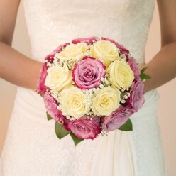 The scene draws you in: a bride stands beneath Queensbury's gentle daylight, holding at her waist an artfully round bridal bouquet brimming with careful intention and artistry. The roses, selected by a local expert and arranged with the devotion one might find at a cherished family celebration on Honeypot Lane, blend soft, creamy yellows with exuberant fuchsia pinks, some petals tinted with understated lavender hues. Petite, airy sprigs of baby's breath, almost cloudlike, weave among the clustered roses, their texture lending an ephemeral grace that echoes the serenity of nearby Canons Park. Tucked amidst the blooms, deep green leaves lend depth and anchor the mix, subtle but essential. The bouquet's fresh, delicate fragrance seems almost tangible, illuminated by even, natural lighting that heightens each detail: the shine of petals, the filigree of lace across the bodice of the bride's classic dress, and the flowing ivory fabric that speaks of timeless traditions. The composition directs all focus to this radiant bouquet-a memory-in-the-making-while the softened background blurs hints of a celebratory gathering. This bouquet, ideal for a Queensbury bride seeking both elegance and heart, becomes more than flowers: it is an emblem of love, anticipation, and the cherished artistry that defines every local wedding celebration.
