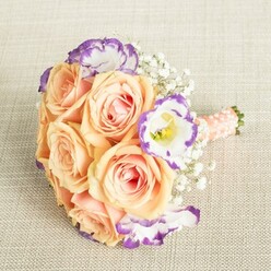 Carrying the gentle spirit of Queensbury, this bouquet is an invitation to linger in the soft glow of celebration. From above, the composition reveals a harmonious cluster of peach and apricot roses, their petals tinged with delicate coral and the faintest blush pink, echoing the warmth of a spring sunrise over Roe Green Park. Interwoven among these classic blooms are lively Lisianthus flowers, crisp white yet edged with whimsical purple, as if each was painted by hand in a moment of inspiration. Threads of Baby's Breath weave throughout-puffs of white that lighten the arrangement, recalling the airy calm along Kenton Lane on a quiet morning. The stems are encased in a wide wrap of peach-toned satin, finished with glimmering pearl beads arranged in subtle order-providing a tangible sense of tradition and refined craftsmanship. This bouquet, round and embracing, would perfectly complement a bridesmaid's hands as she walks into a local parish church or gather the admiration of guests at a family gathering. Every element-texture, colour, fragrance-whispers of careful attention and personal meaning, evoking a celebration rooted in Queensbury's charm. Designed to capture joy and mark life's brightest milestones, it offers a radiant blend of elegance and authentic floral artistry.