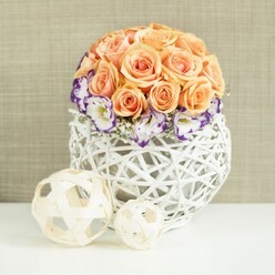 A refined floral centerpiece is presented on a smooth white tabletop, its design balanced and harmonious, much like a thoughtfully laid reception table at a Queensbury hotel or community hall. The arrangement rises in a lush, rounded dome of peach and apricot roses, their petals layered tightly together so that no gaps interrupt the gentle curve. Each rose appears at its peak, with a soft, almost glowing quality that suggests both romance and calm. Nestled around the lower part of the rose sphere, white eustoma blossoms open gracefully, each one edged in a deep, regal purple that frames the rose tones and brings depth to the colour story. Wisps of baby's breath weave between them, their tiny white flowers adding a fine, cloud-like texture. The bouquet is held in a striking bulbous container created from white wicker strands that criss-cross to form an open, lattice-like shell. This woven base feels both rustic and contemporary, the gaps between the strands catching subtle shadows that give the arrangement added dimension. Resting nearby on the same white surface are two light, decorative orbs made from pale wooden strips, one larger and one smaller, echoing the woven pattern of the vase and extending the natural theme. Behind everything, a softly textured beige fabric backdrop creates a neutral, cosy setting, letting the floral colours take centre stage. The entire scene suggests an occasion marked with care and intention, perfect for wedding breakfasts, engagement celebrations, or intimate gatherings in Queensbury homes where a polished yet natural look is desired, and where flowers are chosen as much for atmosphere as for beauty.