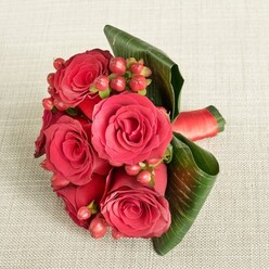 An elegant hand-tied bouquet of rich red roses and berries lies on a pale beige, softly textured fabric, its compact shape and vivid colours creating an immediate sense of romance and warmth. At the center, five or six full-bloom roses open generously, their velvety petals layered in deep crimson tones that catch the light along each curve and edge. Nestled between these roses are clusters of small, rounded Hypericum berries in shades of reddish-pink and soft red, adding lively dots of brightness and a gently bumpy texture that invites the fingertips. Surrounding the blooms, broad, dark green tropical leaves have been artfully wrapped and curled by an experienced florist, forming a sleek, glossy frame that cradles the bouquet and keeps the composition tidy and modern, much like the chic arrangements often requested for weddings and intimate celebrations around Queensbury and the nearby Kingsbury Road. A wide red satin ribbon binds the stems together at the base, its smooth, lustrous surface catching a subtle sheen, suggesting careful craftsmanship and attention to detail from a local workshop such as Flower Delivery Queensbury. The bouquet is photographed from above with soft, even lighting, which accentuates the richness of the red tones and the depth between petals while allowing the neutral background to fade gently out of focus. The overall impression is of a luxurious, romantic arrangement that feels both classic and contemporary, suitable as a bridesmaid's posy, a refined Valentine's gift delivered to a Queensbury doorstep, or a special surprise waiting on a neatly set dining table for an anniversary dinner at home.