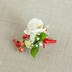 An artfully crafted boutonniere lies centered on a pale oatmeal-coloured woven fabric, its subtle texture reminiscent of the calm, neutral interiors of homes around Queensbury and the surrounding Brent borough. The eye is immediately drawn to a pristine white Phalaenopsis orchid, its broad, smooth petals elegantly curved and slightly overlapping, giving the bloom a serene, sculptural quality. At its core, the orchid reveals a gentle glow of creamy yellow and muted orange, like a tiny sunrise nestled within the flower, adding both warmth and quiet sophistication. Around this focal bloom, delicate baby's breath unfolds in soft clusters of miniature white blossoms, creating a cloud-like halo that lends the arrangement a light, airy feel, perfect for a wedding at a local registry office or a formal celebration in a nearby function room. Alongside the orchid and gypsophila, a small cluster of glossy red berries adds an unexpected burst of colour: the spheres catch the light, appearing almost lacquered, while a few softer green buds promise freshness and natural variety. Slender and broader green leaves weave in and out, offering a verdant frame that balances the bright berries and pale petals. The stems are gathered neatly and wrapped with a deep red satin or silk ribbon, its surface smooth and slightly lustrous, tied into a compact knot that hints at careful, practiced hands. This ribbon tail rests against the neutral fabric, reinforcing the sense of readiness-this is an accessory waiting to be pinned to a lapel for a wedding, winter ball, or prom night somewhere in Queensbury, radiating calm elegance and understated celebration.