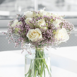 This image focuses closely on a refined sympathy bouquet, where a dozen or more creamy white roses open fully, their petals layered in soft spirals that suggest both purity and tenderness. The roses dominate the arrangement, each bloom unblemished and gently curved, as if cradling the quiet emotion behind the gift. Threaded delicately among them are wisps of lavender and pale pink filler flowers, most likely statice, that create a light, airy veil around the stronger rose shapes. Their tiny clustered petals form a feathery texture, adding depth and dimension without disturbing the bouquet's calm, respectful character. All of the stems gather neatly in a tall cylindrical glass vase filled with fresh, clear water, the green stems straight and vibrant against the glass, a subtle reminder of life and renewal. The arrangement sits on a white surface that reflects just a trace of the vase, while the background dissolves into soft, neutral tones, suggesting a bright, uncluttered space in a Queensbury home or chapel near Honeypot Lane. Gentle, diffused light bathes the bouquet, highlighting the ivory tones in the roses and the cool lavender hues of the filler blooms, creating an atmosphere of tranquillity. One can almost imagine the faint, clean scent of fresh roses, understated and soothing rather than overpowering. The whole composition feels like a quiet companion in times of loss-something a local florist in Queensbury would thoughtfully design to convey sympathy, remembrance, and a touch of peace when delivered to a grieving family.