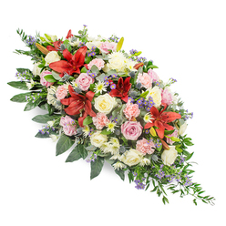 Set against a clean white backdrop, this elongated funeral spray feels like a quiet, floral embrace, the sort of tribute you might see resting on a coffin during a reflective farewell in Queensbury. The design stretches gracefully from a broader, rounded top to a narrower, tapering end, its shape echoing the gentle curve of a final parting. Rich clusters of flowers in shades of deep red, blush pink, creamy white, and soft lavender-purple are packed closely together, creating a sense of fullness and care. Dramatic deep red lilies, with wide, star-shaped petals and velvety markings, form bold focal points through the centre, symbolising strength and devotion. Around them, creamy white and soft pink roses unfold at different stages, from shy buds to open, cupped blooms, suggesting the many chapters of a well-lived life. Light pink carnations add ruffled softness, while petite white chrysanthemums and daisy-like florets slip into the gaps, ensuring no space feels empty or forgotten. Sprigs of spiky blue-purple thistles and tiny lavender-blue fillers peek through the warmer colours, adding cool contrast and a touch of wild, hedgerow character that feels at home in North West London's quieter green corners. The entire spray is cradled in a sweep of lush greenery, with broad, glossy leaves and trailing fern-like fronds that extend beyond the flowers, giving a natural, flowing silhouette. The composition radiates calm remembrance and gentle comfort, the kind of thoughtful arrangement local families in Queensbury might choose for a final goodbye that is both dignified and deeply personal.
