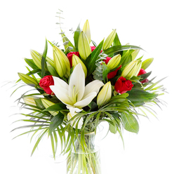 Framed in bright, even light on a stark white backdrop, this floral arrangement reads like a portrait of contrast and careful floral craft - a single, fully open creamy white oriental lily takes centre stage, its smooth, pristine petals curving to expose pale yellow stamens and a pale green pistil that catch the eye. Encircling that central bloom are numerous lily buds in shades of lime green and buttery yellow, some tightly closed, others just unfurling, which gives the bouquet a sense of time and gentle unfolding. Nestled among them, small, velvety red rosebuds offer dramatic punctuation, their concentrated crimson tones suggesting romance without overwhelming the composition. The bouquet is anchored by a variety of greens: glossy dark-green leaves provide a solid backdrop, long, arching palm fronds lend movement and a slightly tropical silhouette, and round, silvered eucalyptus leaves introduce a cool, soft contrast. All of this is arranged in a simple clear glass vase so the light-green stems show through clear water, speaking to freshness and meticulous care. As a local florist have arranged many such pieces for deliveries around Queensbury station, Kingsbury High Road and the edges of Fryent Country Park, I picture the bouquet's fragrance as a blend of powdery lily and warm rose - an aroma that carries through a quiet room or over a celebration table. Flower Delivery Queensbury prepares similar designs for same-day local delivery, perfect for anniversaries, congratulations, or an elegant sympathy tribute.