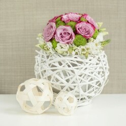 An artfully composed floral arrangement sits serenely on a clean white table, evoking the quiet elegance of a well-dressed Queensbury townhouse set back from the bustle of Kingsbury Road. The centerpiece is a lush, domed bouquet that appears almost cloud-like from above, formed by layers of soft pink and lavender roses with gently ruffled petals. Some blooms are edged in a deeper pink, suggesting the richness of a garden rose at full perfection, and they gather together in a seamless tapestry of romantic colour. Punctuating the roses are bright green button chrysanthemums, their compact, spherical blooms lending structure and lively contrast, like fresh spring grass against the pastel sky. At the base, small white filler flowers weave in and out, adding a feathery border that makes the whole arrangement feel light and breathable. Cradling this floral dome is a round, white woven vase, fashioned from rattan-like branches that curl and interlace to form an open, basket-style sphere. The weaving creates shifting shadows and highlights, emphasised by the soft, even lighting. To the left of the vase, two decorative spheres made of pale, natural wicker rest on the same white surface, one slightly larger than the other, echoing the airy latticework of the container. Behind the scene, a blurred canvas of beige fabric with gentle texture provides a warm, understated backdrop, rather like the neutral interiors of modern apartments near Queensbury Station. The image conveys craftsmanship and calm celebration, perfectly suited to illustrate wedding table flowers, intimate dinner party centerpieces, or stylish home decor ideas for local residents seeking fresh, seasonal blooms delivered across Queensbury.