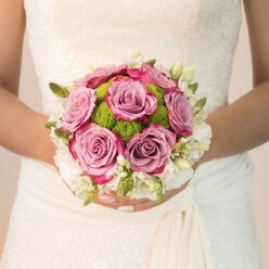 Embodying both artistry and heartfelt romance, this bridal bouquet-photographed as the bride pauses along Kingsbury Road-melds timeless blooms with a burst of contemporary colour. The plush, lavender-pink roses at the center evoke soft, early-morning skies over Queensbury, with each petal touched by threads of deeper fuchsia, as if brushed by the first blush of dawn. Nestled around them, the lime-green pom-pom chrysanthemums lend a playful, textured balance, their fresh hue reminiscent of local gardens in full bloom. The outermost layer, a constellation of delicate, snow-white blossoms and slender green buds, wraps the bouquet in luminosity and classic charm. The bride's ivory gown, detailed with floral lace and subtle draping, adds a touch of vintage sophistication, its craftsmanship mirroring the bouquet's layered finesse. Every element-from the hand-selected flowers to the careful arrangement-speaks to the skill and warmth of Queensbury's local florists. Soft even lighting brings the textures and tints to life, crafting a moment rich with anticipation and joy, ideal for those seeking the perfect wedding bouquet to complement their North West London nuptials. A gentle sense of promise and artistry combines, celebrating love's new beginning amidst the unique backdrop of Queensbury.