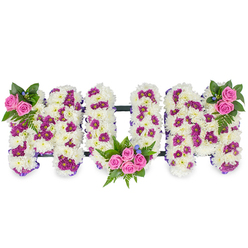 A lovingly composed funeral tribute crafted from thousands of small white chrysanthemum heads, this three-dimensional MUM arrangement reads as both word and soft landscape. The chrysanthemums create an almost cushiony surface, their tightly packed petals producing a serene, luminous white base that feels both delicate and enduring. Interwoven through the white are deliberate accents of deep magenta and purple spray chrysanthemums that give each letter subtle patterning and a gentle rhythm of colour. Bright pink spray roses provide intimate focal points: a petite cluster perched at the upper left of the first M, a more generous bouquet nestled into the lower curve of the U, and a matching cluster crowning the upper right of the final M. Lush fern fronds and varied dark green foliage cradle the roses and lift the shapes, while tiny powder-blue flowers offer small, cool highlights that help the eye move across the composition. A narrow purple ribbon subtly traces the letter edges and a thin, dark horizontal bar links the letters, providing discreet structural unity. The photograph captures soft, diffused light that highlights texture and shadow, enhancing the three-dimensional form. Prepared by a team used to serving Queensbury and the surrounding neighbourhoods, this arrangement can be supplied for chapel services, wakes, or a family memorial; Flower Delivery Queensbury would arrange careful handling and local delivery, ensuring the tribute is presented with dignity and floral expertise.