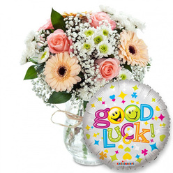 A thoughtful, celebratory arrangement presented with a clean, minimalist backdrop: a clear glass vase cradles a lush mix of pale pink and peach roses, silky to the eye, paired with vibrant peach-orange Gerbera daisies that show off dark brown centers. Interspersed among them are clouds of delicate Gypsophila and small white-and-lime chrysanthemum clusters that add crispness and light, their textures contrasting with the velvet softness of the roses. Deep green foliage threads through the blooms, offering structure and a natural silhouette, while the vase is tied simply with twine for a handcrafted finish. In the foreground, slightly to the right, a shiny silver foil balloon proclaims Good Luck in playful, multi-coloured type; the two o's in Good are bright yellow smiley faces - one winking, one making a cheeky face - and the balloon surface is sprinkled with tiny stars, hearts and colourful geometric dots that catch the light. A discreet Kaleidoscope mark sits on the balloon's lower rim. The whole scene smells faintly of rose perfume and fresh green stems under soft daylight, the petals' edges warmed by gentle highlights. This is the kind of send-off I might prepare for a neighbour heading to Queensbury Tube or a student leaving for an exam near Honeypot Lane - a small, reassuring ritual of colour and scent that translates support into something you can hold.