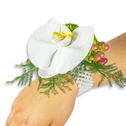 Nestled against the fair wrist of the wearer, a single, luminous white Phalaenopsis orchid serves as the centerpiece of a refined wrist corsage, the image composed against a pristine white backdrop so every texture reads clearly. The orchid's broad, satiny petals open like a small moon, and its central labellum glows with a warm yellow tone that deepens to a golden-orange at the throat, a delicate point of colour in an otherwise white palette. Surrounding the bloom, feathery asparagus fern creates a cushion of bright, spring-green threads that seem to float and tremble with the slightest movement. Close by, a cluster of Hypericum berries gives the eye a quiet punctuation: glossy spheres that graduate from pale green to soft rose and finally to a rich cranberry, their sheen like tiny polished beads. Interspersed lime-green accent leaves add a subtle ruffle and lift the composition's silhouette. The corsage is secured on a classic bracelet of tiny white pearls, multi-stranded and gently yielding to the wrist - a tactile heirloom detail that pairs modern freshness with timeless refinement. Under neutral studio light the materials read clean and true: smooth orchid skin, the papery firmness of the berries, the soft give of the fern. Emotionally, the piece whispers of first dances, thoughtful vows and small public moments of pride; it is the kind of wearable bloom a Queensbury family might choose for prom night, a wedding in a local church, or a community celebration near Kingsbury and Wembley. The overall impression is calm, elegant and expertly crafted by hands that respect both flower and wearer.