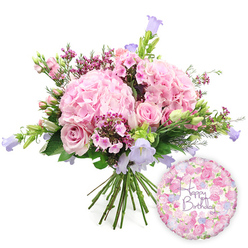 A lush, celebratory birthday bouquet is set against a crisp white backdrop, allowing every pink and purple petal to stand out clearly, much like a shopfront display along Queensbury's main parade on a sunny afternoon. The arrangement is hand-tied and deliberately full, with light blush pink hydrangeas forming generous, cloud-like mounds in the centre. Their ruffled petals overlap in soft layers, creating a romantic, tactile texture that feels both luxurious and comforting. Woven among these hydrangeas are tender pink roses, some fully open with their swirling centres on show, others held in tighter buds that suggest the flowers will continue to unfold over the coming days. Clusters of deeper fuchsia filler flowers, similar to waxflower, nestle in between, delivering tiny star-shaped blooms and adding a playful contrast to the gentler tones. Tall, graceful stems of campanula rise through the composition, their bell-shaped flowers in pale lavender and periwinkle blue lifting the eye upwards and bringing a cool, calming note to the palette. A variety of fresh foliage, from pointed green leaves to softer, rounded greens, fills out the bouquet, giving it an organic, garden-inspired feel that would look perfectly at home in a Queensbury living room after a doorstep delivery. The stems are visible and neatly gathered at the bottom, confirming the professional hand-tied craftsmanship. Resting to the lower right side of the bouquet is a shiny round foil balloon, its surface adorned with a delicate floral pattern reminiscent of miniature rosebuds and soft pastel blooms. At its centre, the words "Happy Birthday" are written in elegant purple script, tying the entire scene together as a joyful birthday gift that combines flowers and balloon in one thoughtful gesture for someone special in Queensbury.