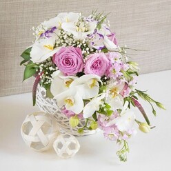 An elegant wedding-style table arrangement is captured in a close, carefully composed photograph, reminiscent of a beautifully laid reception table at a cosy venue in Queensbury. The centerpiece is an abundant bouquet cradled in a round white wicker basket, the weave crisp and tidy, yet rustic enough to feel handcrafted. From this basket spill generous blooms of pastel pink roses, fully opened so that their spiral hearts are clearly visible, each petal curving softly outward with a velvety sheen. Intertwined with the roses are pristine white Phalaenopsis orchids, their broad, butterfly-like petals framing golden-yellow centers touched with tiny hints of orange and purple, adding a touch of exotic luxury. Around them, ruffled white Lisianthus flowers, some edged with a whisper of lavender, bring softness and depth, while stems of pale pink stock rise and gently arch, suggesting height and a light, powdery fragrance. Airy white baby's breath is scattered through the bouquet like fine lace, filling in gaps and giving the arrangement a cloud-like fullness. Fresh green leaves and a few tender buds peek out between blossoms, breaking up the pastels with natural, vibrant contrast. Resting on the white tabletop in front of the basket are two decorative orbs made from thin, interlaced natural fibers, one slightly larger than the other, echoing the wicker texture and adding a sculptural, minimalist touch. Behind the flowers, a neutral beige or taupe fabric backdrop, similar to soft linen, provides a gentle, unfussy canvas so the colours appear bright yet soothing. The scene feels ideal for a Queensbury wedding breakfast or an anniversary dinner at home, the kind of refined yet welcoming design a local florist might deliver to families along Honeypot Lane or near Kingsbury Circle.