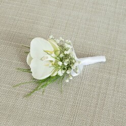 A single white orchid boutonniere lies gracefully on a pale beige, woven fabric surface, its texture reminiscent of natural linen you might find in a cosy Queensbury café overlooking the busy main road. The boutonniere's focal point is a pristine Phalaenopsis orchid, its large, velvety petals spreading out in a serene, symmetrical form. The orchid's throat reveals a soft wash of pale yellow, like a hint of early morning light, giving the bloom depth and a sense of quiet warmth. Nestled around the orchid are airy clusters of baby's breath; each tiny Gypsophila flower forms a delicate constellation of white, creating a gentle, cloud-like frame that makes the central blossom appear even more refined. Wisps of fresh green foliage, likely fine fern or similarly delicate greenery, weave in and out of the white flowers, their subtle curves adding movement and a natural, garden-fresh feel. The stems are meticulously gathered and bound with a smooth white satin ribbon, wrapped snugly and finished with a small, precise bow, indicating the piece is ready to be pinned to a lapel. Soft, diffuse lighting enhances the natural tones of cream, white, and green, and brings out the tactile quality of both the ribbon and the background fabric. The scene suggests the quiet moment before a ceremony, perhaps in a Queensbury registry office or a nearby church, when the groom or groomsman has this elegant detail pinned in place. It conveys a sense of romance, care, and understated luxury, the sort of handcrafted finish local florists create when designing wedding flowers to be delivered fresh across Queensbury and the surrounding neighbourhoods.