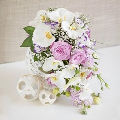 On a smooth white tabletop, a luxurious floral centerpiece glows softly against a neutral beige wall, as if prepared for an intimate celebration in a quiet Queensbury dining room. At its heart stands a white, spherical vase fashioned from an intricate weave of slender strands, reminiscent of delicate wicker or a carefully wrapped ball of twine. From this contemporary base springs a lush dome of flowers in shades of white, blush, and lilac. Large Phalaenopsis orchids in pure white open wide, their petals broad and serene, while their inner throats shimmer with golden-yellow and soft amber, adding an exotic accent that draws the gaze. Nestled around them are plump roses in pastel pink and lavender, some fully open to reveal every layered petal, others just beginning to loosen, giving a sense of living, unfolding beauty. Between these, smaller ruffled blooms, likely Lisianthus or Eustoma edged in purple, introduce texture and a painterly quality. Wisps of white baby's breath drift through the bouquet like tiny stars, filling gaps and creating a cloud-like softness. Slender stems of pale lilac stock and trailing blossoms spill gently over the rim of the vase, suggesting a natural, flowing garden rather than something rigidly formal, while rich green leaves add depth and contrast. At the side, two decorative open-weave balls in a creamy white echo the vase's texture and hint at thoughtful styling, perfect for a wedding table along Honeypot Lane or a refined anniversary dinner near Queensbury Park. The overall mood is soothing, elegant, and quietly celebratory, as if the flowers have been freshly arranged for same-day delivery by a florist who understands both design and the relaxed rhythm of local life.