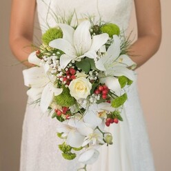 As the bride stands in the gentle light filtering through the windows near Queensbury's peaceful residential streets, her hands hold an abundant cascading bouquet-a living tapestry that combines old-world charm with contemporary flourish. The eye is first drawn to majestic white lilies, their large, ruffled petals painted with subtle cinnamon speckles, filling the bouquet with a pure, luminous energy. At the heart, a classic cream rose unfolds in smooth, velvety layers, its scent whispering a promise as timeless as the vows soon to be spoken. Dotted throughout are pops of lime green cushion mums, whose playful texture and colour reflect the vibrancy found in local gardens and sunny parks nearby. Wispy sprays of baby's breath float like tiny clouds, bringing lightness and breathing space between the structured blooms. For a joyful seasonal accent, clusters of bright red berries glisten among the petals, adding a delightful contrast reminiscent of festive holidays shared with family. The bouquet's signature cascade is created by arching stems of elegant white Phalaenopsis orchids that trail downward, their blooms catching every ray. Surrounding greenery and trailing grass blades form a frame, their lively tones reminiscent of the lush greens along the nearby Canon Lane. The bride's graceful hands and the delicate lacework of her gown complete the scene, making this bouquet a perfect inspiration for couple's seeking sophisticated, meaningful wedding flowers in Queensbury.