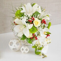 On a pristine white tabletop, a lavish floral arrangement in a rounded white wicker basket creates a striking focal point, the kind of centrepiece you might find gracing a wedding reception in Queensbury or a smart dinner gathering off Kenton Road. The basket's woven texture is echoed by two decorative white wicker spheres resting to the left, their open latticework catching the soft, neutral light against a gentle beige fabric background. From the basket rises a sculpted mass of white Oriental lilies, their wide trumpet blooms unfurling with visible speckles and prominent stamens, suggesting a subtle, powdery scent. Near the upper centre, a single cream-yellow rose, still slightly budded, adds a buttery glow, anchoring the eye amid the surrounding white. Sprays of delicate baby's breath weave between the main flowers, like tiny white stars, while bright green pom-pom blooms and tufts of mossy dianthus lend a fresh, contemporary twist. Wisps of fine greenery and feathery foliage spill outward, the mix of textures hinting at the experienced hand that placed every stem. Along the right edge of the design, white phalaenopsis orchids cascade down in a graceful waterfall, their petals smooth and luminous, creating a soft sense of movement. Scattered here and there are clusters of glossy red berries, providing rich crimson accents that feel festive without overpowering the composition. The overall mood is one of pure, understated elegance and gentle celebration, a refined floral statement that Flower Delivery Queensbury could easily imagine on a bridal table or special family gathering, delivered fresh the same day to homes and venues across the area.