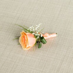 A softly luminous peach rose boutonniere is displayed against a pale beige, linen-textured background, the fabric's fine weave forming a quiet, neutral canvas that allows every petal and leaf to stand out. The rose itself is slightly off-centre and positioned horizontally, its partially opened bloom revealing a heart of warm, sunset peach that slowly fades into creamy, almost shell-like tones at the edges. Nestled to one side, delicate sprays of white baby's breath cluster like tiny snowflakes, their airy forms adding a whisper-light, romantic detail that contrasts beautifully with the rose's smoother, fuller curves. From behind the flowers, slim strands of vibrant green foliage stretch and peek through, while two broader, darker green leaves are gently curled around the base of the stems, giving the design both visual depth and subtle support. The stems are expertly wrapped with a coordinating peach satin ribbon, its surface catching soft highlights and ending in a compact knot that suggests readiness for pinning onto a suit jacket or prom dress. The lighting is gentle and even, reminiscent of an overcast yet bright day in Queensbury, perhaps near the green spaces where many couples head for wedding photos, creating minimal shadows and emphasizing the velvety texture of the rose and the dainty detail of the gypsophila. The composition conveys a mood of quiet elegance and heartfelt celebration, the kind of refined floral touch a local florist would prepare for weddings, proms, or intimate ceremonies across the neighbourhood.