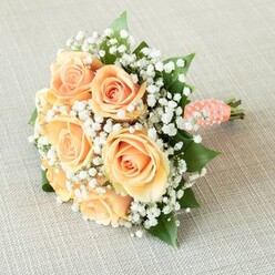Captured against a pillow of soft beige linen, this hand-tied bouquet tells a story of gentle romance and meticulous craftsmanship, as beloved throughout Queensbury as the Sunday strolls down Church Lane. At its heart, approximately eight glorious roses unfold their petals in shades of peach and pale apricot, each bloom brimming with the kind of quiet promise that marks life's special celebrations. Feather-light sprays of pristine white gypsophila curve around the roses, lending a sense of lightness and purity, much like morning clouds skimming over Roe Green Park. Vibrant emerald leaves shimmer from beneath, complementing the pastel palette and grounding it in nature's rich hues. The bouquet is shaped into a soft, rounded arrangement-reminiscent of classic wedding traditions cherished by local families. Binding the stems, a smooth satin ribbon in warm coral-peach sweeps around the handle, its surface embellished with pearly white beads spiraling in a delicate motif, echoing the subtle luxury of vintage bridal accessories. The bouquet evokes the tender fragrance of fresh blooms mingled with green stems, conjuring a sense of joyful anticipation and heartfelt celebration. Illuminated with even, natural light, each detail is brought to life-an arrangement thoughtfully crafted by Queensbury's experienced florists, perfect for bridal parties, anniversary gifts, or memorable gatherings held close to home.