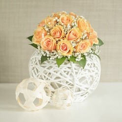 A lush, domed bouquet of peach roses and white baby's breath sits gracefully on a pristine white tabletop, evoking the feeling of a carefully prepared celebration in a quiet Queensbury townhouse. Around twenty to thirty roses, in shades ranging from soft apricot to warm honeyed peach, are tightly clustered in a rounded mound, their petals layered like delicate silk and slightly ruffled at the edges. Deeper orange tones glow from the centres of the blooms, while the outer petals soften to pale, creamy peach, giving the arrangement a warm, candlelit radiance even in daylight. Threaded between the roses are fine stems of white gypsophila, each tiny flower forming part of a cloud-like ring that adds lightness and texture. Here and there, rich green leaves provide a refreshing contrast, echoing the feel of a well-tended garden near Queensbury Park. The flowers are presented in a large, spherical white container crafted from interwoven strips that form an open, airy lattice; it has the look of carefully woven rattan or wicker, modern yet natural. In front of this main vessel sit two smaller decorative spheres in soft beige and cream, their open weave and geometric lines echoing the texture of the vase and adding a thoughtful design detail. Behind the arrangement, a subtly textured light beige wall creates a quiet, neutral backdrop, allowing the colours to stand out without distraction. Soft, even light fills the scene, gently highlighting each petal and casting delicate shadows from the woven elements, suggesting an atmosphere perfect for an intimate dinner, a wedding reception, or a stylish home interior in Queensbury.