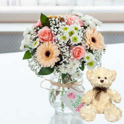 On a clean white tabletop, a romantic anniversary scene unfolds in soft, bright natural light, reminiscent of a calm afternoon near Queensbury's quiet residential streets. A clear glass jar-style vase holds a carefully designed bouquet, where every stem seems thoughtfully placed. Blush pink roses open gently beside peach gerbera daisies, their dark brown centres providing a striking, almost chocolatey contrast. Between them, fine sprays of white baby's breath weave in and out like lace, while small white chrysanthemums with hints of lime-green at their centres lend a cool, refreshing brightness. A length of natural twine is wrapped around the neck of the vase and tied in a rustic bow, from which a small white tag dangles, printed with a simple pink heart and the word Handmade in fluid green lettering, suggesting a gift prepared with care rather than something rushed. To the right of this floral display, a cream-coloured teddy bear sits upright, body slightly leaned toward the bouquet as if sharing the moment. Its fur looks soft and fluffy to the touch, with tiny dark eyes, a brown button nose, and a slim, neatly tied brown satin ribbon around its neck that hints at old-fashioned charm. The white surface beneath them catches a faint reflection of both bear and flowers, while the blurred background suggests a bright window or airy hallway in a Queensbury home. Together, the bouquet and teddy bear create a picture of affection and comfort, perfect for celebrating an anniversary, a first date remembered, or a quiet I love you sent with local flower delivery across Queensbury.