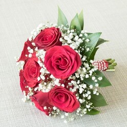 Artfully arranged by a skilled local florist, this radiant bouquet pays homage to the enduring beauty so often seen in Queensbury's vibrant celebrations. Rich, velvety red roses-five or six in full, exuberant bloom-take centre stage, their tightly furled petals expanding into luminous whorls of deep crimson. These iconic roses, symbols of romance and devotion, are softened by generous clouds of delicate white Gypsophila, their miniature blossoms floating like gentle spring mist above the structured greenery. The bouquet gains depth and brightness from lush, dark green leaves, possibly ruscus or salal, peeking between each bloom and providing a natural, refreshing contrast. At the stems' base, the hand-tied bouquet is meticulously finished with a bold red and white striped ribbon, accented with a slender string of tiny, pearly beads that glint in the light-details that speak to a florists' careful artistry often found at Flower Delivery Queensbury. Gently resting atop a pale, textured fabric-perhaps a familiar sight in the cosy homes of Honeypot Lane-the arrangement is softly illuminated, inviting warm feelings of intimacy, celebration, and gratitude. Whether for a wedding along the canal, an anniversary along Colindale's tree-lined avenues, or a spontaneous romantic gesture, this bouquet encapsulates the freshness and heartfelt sentiment that Queensbury locals cherish.
