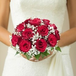 A close, loving embrace between hands and flowers, this stunning Queensbury bridal bouquet encapsulates the spirit of weddings celebrated along the leafy lanes and vibrant squares of the area. Centred around a lush cluster of velvety red roses in full, passionate bloom, the bouquet's domed shape and intricate layering reveal a florist's flair for harmony and detail. Each rose is tightly furled yet inviting, exuding a subtle, sweet fragrance that carries echoes of bouquets chosen on special mornings throughout Queensbury's lively market streets. Ethereal sprays of crisp white baby's breath weave their way through the bouquet, their delicate presence softening the arrangement's bold hues and lending a touch of classic romance reminiscent of local garden gatherings. From the bouquet's base, a fresh hint of green foliage emerges, adding a lively counterpoint and reminding one of the lush serenity found in the parks just beyond Kingsbury Circle. Gently cradled by hands with perfectly polished nails, the bouquet is set against the gentle texture of an embroidered or lace-trimmed wedding dress, its fine details shimmering in soft daylight that feels both modern and timeless. The background dissolves to a hushed, warm neutral, inviting quiet focus on the heartfelt union of bride and blooms. Ideal for those planning weddings in Queensbury, this composition speaks of artistry, devotion, and the gentle beauty inherent in every matrimonial promise.