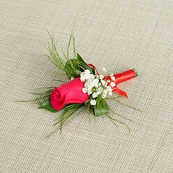 An elegant single-stem boutonnière is captured in bright, natural light, resting gently on a pale beige, linen-like fabric that echoes the soft, neutral décor often used in Queensbury wedding halls and local event spaces. The focal point is a tightly closed rosebud in a vivid fuchsia to deep magenta shade, its petals richly saturated and velvety, forming a compact swirl of colour that feels both romantic and modern. Encircling this central rose, clusters of delicate white baby's breath form a fine, airy halo, each tiny bloom contrasting crisply against the darker petals and adding a sense of softness, like a light mist around a sunset sky. Behind the flowers, fine, bright green foliage extends outward in slender, feathery strands, mingling with a few slightly broader leaves to create depth and a fresh, just-picked-from-the-garden look, as though it were gathered from a well-tended plot near local Queensbury homes. The lower stems of the rose and greenery are carefully aligned and bound together with a smooth, coral-leaning red satin ribbon, neatly wrapped to create a slim, glossy handle that catches the light with subtle reflections. Every detail in the image speaks of skilled craftsmanship and attention to proportion, from the gentle fan of greenery to the precise balance between bloom and filler. The composition evokes the excitement of a wedding morning or prom night, when small floral touches make a suit feel complete, and could easily be imagined gracing a lapel on the way to a celebration in or around Queensbury, prepared with the kind of local care guests appreciate on such meaningful occasions.
