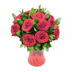 A classic, round bouquet of approximately twelve to fifteen deep crimson roses sits confidently in a red glass vase, photographed against a pristine white backdrop that makes every detail of the arrangement stand out. Each rose is at its prime, with velvety petals unfolding in perfect spirals that reveal the heart of the bloom without appearing overblown or tired. The petals curve and overlap like soft satin ribbons, building a dense, cushion-like surface of rich, ruby colour that immediately suggests romance and devotion. Around and between the roses, dark green foliage-most likely ruscus-extends in smooth, glossy leaves, their pointed shapes and polished surfaces providing structure and contrast, as though gently cradling the blooms. The vase is made of translucent red glass with a softly flared neck and a curving silhouette that narrows gracefully towards the base, its subtle ribbed texture catching glimmers of light and shifting tones from deep burgundy to glowing cherry. Through the glass, the stems are only faintly visible, giving an impression of depth and grounding without distracting from the flowers. The bright, even lighting eliminates distractions, so the focus stays on the interplay of red and green and the subtle reflections in the vase. It feels like the kind of luxurious bouquet a local Queensbury florist would prepare for a milestone anniversary or a Valentine's Day delivery to a home near the station, where only red roses will do. There is an air of sincerity and intensity here, a floral statement that quietly says, "You matter," in the language of roses.