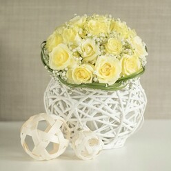 This image shows a beautifully composed wedding table centerpiece that could easily grace a reception in one of Queensbury's intimate function rooms or a quiet home celebration just off Honeypot Lane. The eye is immediately drawn to a generous, dome-shaped bouquet of light yellow roses, their colour reminiscent of sweet cream or early morning sunshine. Each rose head is densely packed, with plush petals curling outward in soft, concentric layers, creating a feeling of fullness and quiet luxury. Threaded between the roses are tiny white blooms of baby's breath, forming fluffy, cloud-like clusters that add a light, romantic haze and a slightly wild, garden-inspired charm.

At the base of the bouquet, a thin band of glossy green foliage is wrapped neatly around the stems, its smooth surface and cool tone providing a crisp contrast to the buttery flowers and gauzy gypsophila. The arrangement is set into a distinctive white woven vase shaped like a rounded basket, its open, lattice-like structure made from interlaced strands that give it the look of modern rattan. Light filters through this pattern, creating intricate shadows on the pristine white table surface below. On the tabletop, two natural-looking woven balls in pale wood tones rest casually beside the vase, repeating the same openwork motif and giving the scene a cohesive, thoughtfully styled feel.

The background is a soft, blurred wash of warm beige, similar to the painted walls of many Queensbury homes, ensuring all attention remains on the floral artistry. The overall impression is one of gentle joy and understated sophistication, perfectly suited to wedding breakfasts, engagement dinners, or relaxed yet elegant gatherings across Queensbury and nearby Harrow.

