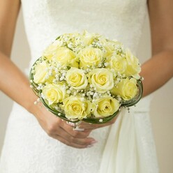 Bathed in soft indoor light reminiscent of a Queensbury morning, this lavish bridal bouquet embodies quiet luxury and timeless tradition. Shapely, pale yellow roses crowd together, their petals curling gently in harmonious whorls, forming a plush, spherical cluster alive with subtle gradients of lemon and cream. Interspersed between the roses, clouds of ethereal baby's breath shine with clean, snowy contrast, infusing the bouquet with a sense of levity-almost like a whisper of blossom-laden air drifting over Kingsbury Road. A refined border of deep green leaves or decorative wire encircles the base, creating a sense of polished structure while recalling the lush greenery of local gardens. At the heart of the moment, hands with softly manicured nails and a gleaming ring delicately support the bouquet, their posture heavy with meaning and poised expectation. The rich texture of a white wedding gown provides a canvas of subtle embroidery and dreamlike softness in the background, with bright, natural light enhancing every petal's sheen. For both brides and flower lovers across Queensbury, this arrangement captures not just the artistry of local floristry but the enduring hope and joy that celebrate the first day of a new journey together. It is the kind of bouquet that promises both fragrance and memory-crafted with care for those who value romance, style, and thoughtful detail.