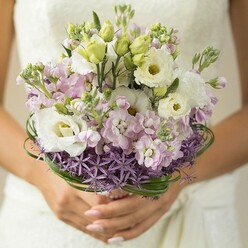 Bathed in soft daylight reminiscent of Queensbury's tranquil mornings, this bridal bouquet evokes a calm, radiant happiness that lingers in the air. Within the gentle embrace of a bride's hands, delicately manicured in a pale nude hue, the bouquet becomes a centerpiece of artistry and emotion. Creamy white lisianthus open with vintage elegance, their papery petals catching the light just so, while slender, unopened buds stand quietly beside them, promising future blooms and shared memories. Fragrant sprays of pale lavender and light pink stock flowers spill between the lisianthus, their layered textures creating both visual and aromatic harmony-a nod to the fresh florals often sourced from nearby local markets. The base glimmers with the unique, architectural beauty of purple allium, its star-shaped blossoms introducing a playful geometry under the softer forms. Bands of glossy bear grass spiral gracefully, lending a modern structure that reflects both intention and spontaneity. In the background, a subtle lace or ivory gown softly blurs into the composition, evoking a sense of timeless celebration. This arrangement-thoughtfully designed for a joyful day in Queensbury-radiates hope, pure affection, and the heartfelt skill that local florists bring to each wedding. The bouquet's scent gently dances on the breeze, an echo of spring mornings walking through the neighborhood's leafy avenues.