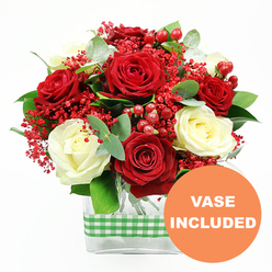 A rich, romantic bouquet of red and white roses is shown in crisp detail against a pure white backdrop, as if waiting to be placed on a dining table in a Queensbury townhouse. The arrangement is dominated by full, velvety red rose heads, their petals layered in loose spirals that suggest passion and warmth, balanced by softly glowing cream and ivory-white roses that bring a serene, elegant note. Between these classic blooms, tiny red filler flowers cluster like a cloud of sparkling confetti, their fine stems branching in all directions to create a light, airy halo of colour. Round, polished red hypericum berries punctuate the design, adding little globes of shine that echo the red roses but with a firmer, more structured form. Lush green foliage weaves through the bouquet: broad, glossy leaves provide a sturdy backdrop, while the muted, silvery-green eucalyptus leaves add movement and a contemporary twist, like greenery freshly cut from gardens off Honeypot Lane. The stems are gathered into a sleek, rectangular clear glass vase, where the clean lines of the container contrast beautifully with the softness of the petals. Around the centre of the vase, a green and white gingham ribbon is snugly wrapped, its cheerful check pattern lending a homely, country-kitchen charm that feels at once stylish and familiar. In the bottom right corner of the image, an orange circular label is overlaid with the words VASE INCLUDED in white capital letters, making it clear that this floral gift arrives complete and ready to display, perfect for same-day delivery in Queensbury when you want to express love, gratitude, or festive cheer without any extra fuss.