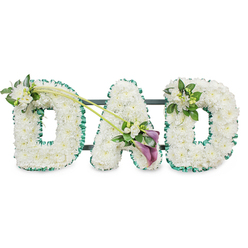 The image shows an elegant "DAD" funeral tribute, created as three bold, three-dimensional letters that stand side by side, all formed from masses of tightly arranged white chrysanthemums. Each tiny floret is fresh and full, combining to form a plush, cloud-like surface that suggests purity, peace, and a gentle final goodbye. Along the outer edge of every letter runs a rich, emerald green ribbon, slightly ruffled and satin-soft, which gives a clear outline and infuses the design with a touch of natural vibrancy. Behind the flowers, you can just make out slim dark bars that reveal a sturdy frame, allowing the piece to stand upright at a funeral service in Queensbury, perhaps at a chapel on Church Lane or in a quiet crematorium setting. The left-hand "D" carries a small, graceful posy of white freesia and lush foliage on its upper curve, the blossoms lifting the line of the letter with a subtle, hopeful air. Two slender green stems sweep diagonally from this first "D" towards the centre, connecting visually with the "A" and guiding the gaze along the tribute. Where these stems meet the base of the "A", a deeper accent draws attention: two rich, purple calla lilies, with their smooth, sculpted trumpets, nestle among more white freesia and glossy greenery, creating a focal point that feels both modern and timeless. The final "D" is finished with a smaller cluster of freesia and foliage on its upper right, echoing the other side and bringing balance. Overall, it is a lovingly crafted memorial piece, ideal for a Queensbury family wishing to honour a father with dignity and heartfelt remembrance.