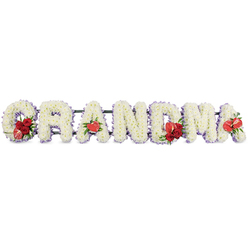 The image shows a beautifully composed funeral flower tribute spelling "GRANDMA" in large, upright capital letters, arranged in a straight line as if ready to be placed beside a coffin or at the front of a chapel in Queensbury. Each individual letter is built from tightly massed white chrysanthemums, their neat, button-like blooms pressed closely together to form a dense, cushiony surface with a soft, almost quilted appearance. Around the outside edge of every letter runs a slim band of pale lavender or light purple ribbon, giving the design gentle definition and adding a faint, soothing wash of colour that softens the starkness of the white. At key points along the lettering, rich red flowers introduce warmth and emotion: the "G" features a luxurious cluster of deep red roses combined with an eye-catching red and white variegated calla lily, tucked into the lower curve. The first "A" carries a single, shiny, heart-shaped red anthurium, positioned at its base like a small emblem of love. In the centre, the "M" is decorated with another group of red roses, surrounded by lush green foliage and partnered with a matching red anthurium. The final "A" is finished with its own solitary anthurium bloom, echoing the earlier accents and bringing balance to the piece. A dark green metal or plastic stand supports the letters just behind, keeping them aligned while remaining visually subtle. The tribute feels thoughtful and tender, the kind of bespoke arrangement a Queensbury family might choose from a trusted local florist to remember their grandmother with dignity and affection.
