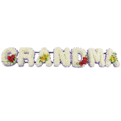 The image shows a lovingly crafted GRANDMA funeral flower tribute, laid out in strong capital letters as if resting gently in the air against a crisp white background. Each letter is carefully formed from masses of small white chrysanthemums, packed closely together so that the surface feels plush, soft, and completely covered in petals, with no gaps or greenery showing through. Beneath this snowy top layer, a subtle band of pale lavender blooms peeks out, outlining every curve and corner and casting a faint shadow of colour that helps each letter stand proud and readable, even from a distance in a church or crematorium chapel near Queensbury. The arrangement is thoughtfully accented with a few bolder flowers to personalise the piece. At the lower curve of the G, three luxurious, deep red roses open towards the viewer, surrounded by fresh dark leaves that provide contrast and depth. The first A is gently embellished with two yellow-green orchids, their unusual colour and speckled reddish centres lending a touch of individuality that might echo a grandmother's favourite shade or treasured garden plant. The central R, N, and D remain entirely white and lavender, like a calm pause in the middle of the tribute. On the right-hand side, the M holds another trio of red roses in its lower arc, while the final A repeats the pairing of chartreuse orchids, bringing the design full circle. A slim, dark framework discreetly connects the letters at the back, ensuring the tribute is secure for local delivery and display, while the overall piece radiates quiet love, remembrance, and gratitude for a cherished matriarch.