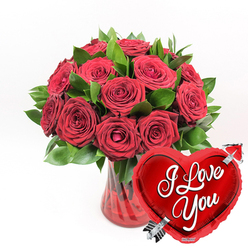 A richly romantic bouquet of deep red roses commands attention at the centre of a bright white background, every blossom carefully composed to celebrate love in its most classic form. Around a dozen or more roses, all in varying stages of bloom, are clustered together so closely that their velvety petals seem to spill over each other, creating a soft, luxurious cushion of crimson. Some heads are fully opened, revealing spiralled centres with subtle shadows in their folds, while others remain partly closed, their edges still tightly rolled like whispered promises. Threaded between the roses, lush green foliage adds structure and freshness, the cool emerald tones providing a beautiful contrast to the warm reds. All of this is gathered into a clear, ruby-red glass vase with a gentle fluted texture, the ridged sides catching and diffusing the light so the vase glows like a precious gem set on a white canvas. The stem ends and water are faintly tinted by the red glass, intensifying the bouquet's romantic atmosphere. Just in front, to the lower right, a glossy red heart-shaped foil balloon leans towards the flowers as if in conversation. Its reflective surface shows crisp highlights, and across the centre the words I Love You appear in flowing white script. A silver Cupid's arrow seems to pass diagonally through the heart, adding a playful nod to old-fashioned courtship. This image feels like the kind of grand, heartfelt gesture a Queensbury local might send through Flower Delivery Queensbury to mark an anniversary, Valentine's surprise, or any day when a simple message of love deserves something unforgettable.