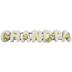 A carefully crafted floral tribute spells "GRANDPA" in bold, upright letters, set horizontally as though resting gently before a funeral service in Queensbury. Each of the seven uppercase characters is densely packed with pristine white spray chrysanthemums, their countless petals forming a soft, cushiony surface that looks almost velvety to the touch. Hints of pale yellow at some flower centres lend a natural warmth, while the overall white expanse speaks of respect, remembrance, and a peaceful passing. Tracing the outline of every letter is a slim band of light lavender or lilac blooms, which create a gentle contrast, sharpening the silhouettes of the G, R, A, N, D, P, and final A against the stark white background. This lilac edge has the quiet calm of a soft evening sky above the rooftops near Queensbury Station.   

Several letters are embellished with small, harmonious sprays of accent flowers. The "G" features a prominent grouping of three to four peach-toned roses, some fully open, others still cupped in bud, combined with narrow white lily buds and polished green leaves. On the first "A", a single peach rose with surrounding foliage is tucked into the flowers, adding a personal, intimate touch. The "D" holds another cluster of peach roses and greenery, reflecting the styling of the "G" to bring balance across the word. The closing "A" is finished with one more peach rose and fresh leaves, like a final whispered farewell. A thin, dark bar runs along the base, hinting at a discreet metal or foam frame that supports the entire arrangement, ready to be displayed at a local chapel or graveside. This personalised GRANDPA tribute feels both strong and tender, a floral message of love created for a beloved grandfather and the family who will remember him.