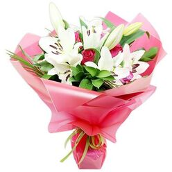 The image presents a thoughtfully crafted bouquet that marries the purity of white Oriental lilies with the passionate depth of red roses, styled with the calm precision of an experienced Queensbury florist. The lilies stand tall and generous, their long, structured petals opening into star-shaped trumpets that reveal warm, chestnut-coloured stamens and slender pistils at the centre. A few lilies remain in tightly sheathed buds, tinged with soft green and creamy white, indicating both freshness and longevity. Between these architectural blooms sit compact, velvety red rosebuds and partially opening roses, each cupped with layers of petals that spiral inward, giving the arrangement an intimate, romantic heartbeat. The flowers emerge from a bed of rich green foliage-sturdy, polished leaves and finer, arching blades that create a sense of natural movement, like a breeze drifting through the tree-lined streets off Stag Lane. All of this is embraced by voluminous, bright pink cellophane wrapping, its glossy, semi-transparent surface folding in generous ripples around the stems. The pink catches the light and reflects softly onto the white petals, lending a gentle blush to the lilies. At the base, a slim lime green ribbon is tied into a small, tidy knot, the ends trailing lightly to soften the finish. The bouquet feels festive yet elegant, equally fitting for a Mother's Day surprise, a heartfelt thank-you to a neighbour in Queensbury, or a romantic gesture delivered the same day to a loved one on a quiet residential street just off the Broadway.
